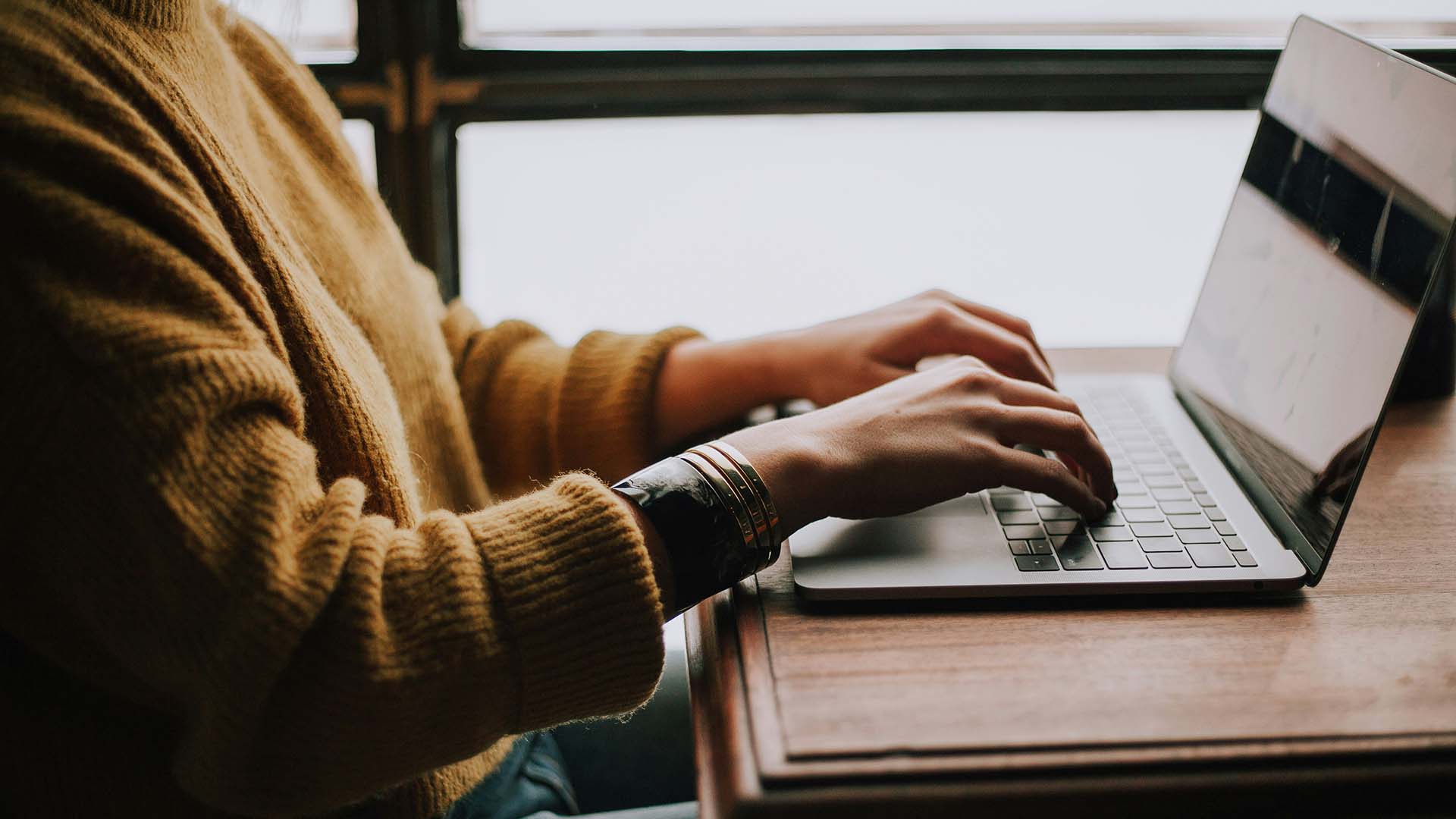 Person in sweater sitting at table working on laptop computer.